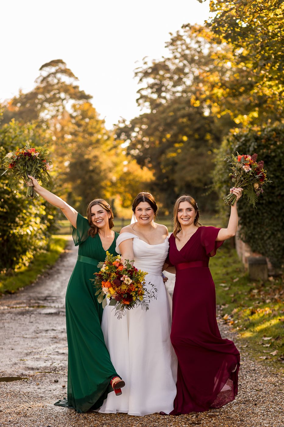 Three people in jewel-toned dresses pose together on a autumn path with colorful bouquets.