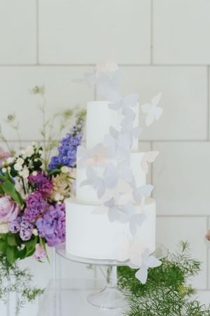Elegant white tiered wedding cake decorated with delicate pastel butterflies beside purple and white floral arrangements.