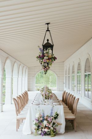 Elegant wedding reception table set with white linens and floral arrangements under a covered porch with arched windows.
