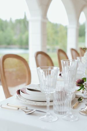 Elegant table setting with crystal glasses, white plates and wooden chairs on a covered porch overlooking greenery.