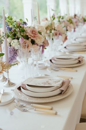 Elegant wedding table setting with blush roses, crystal glassware, and white plates arranged on a pristine tablecloth.