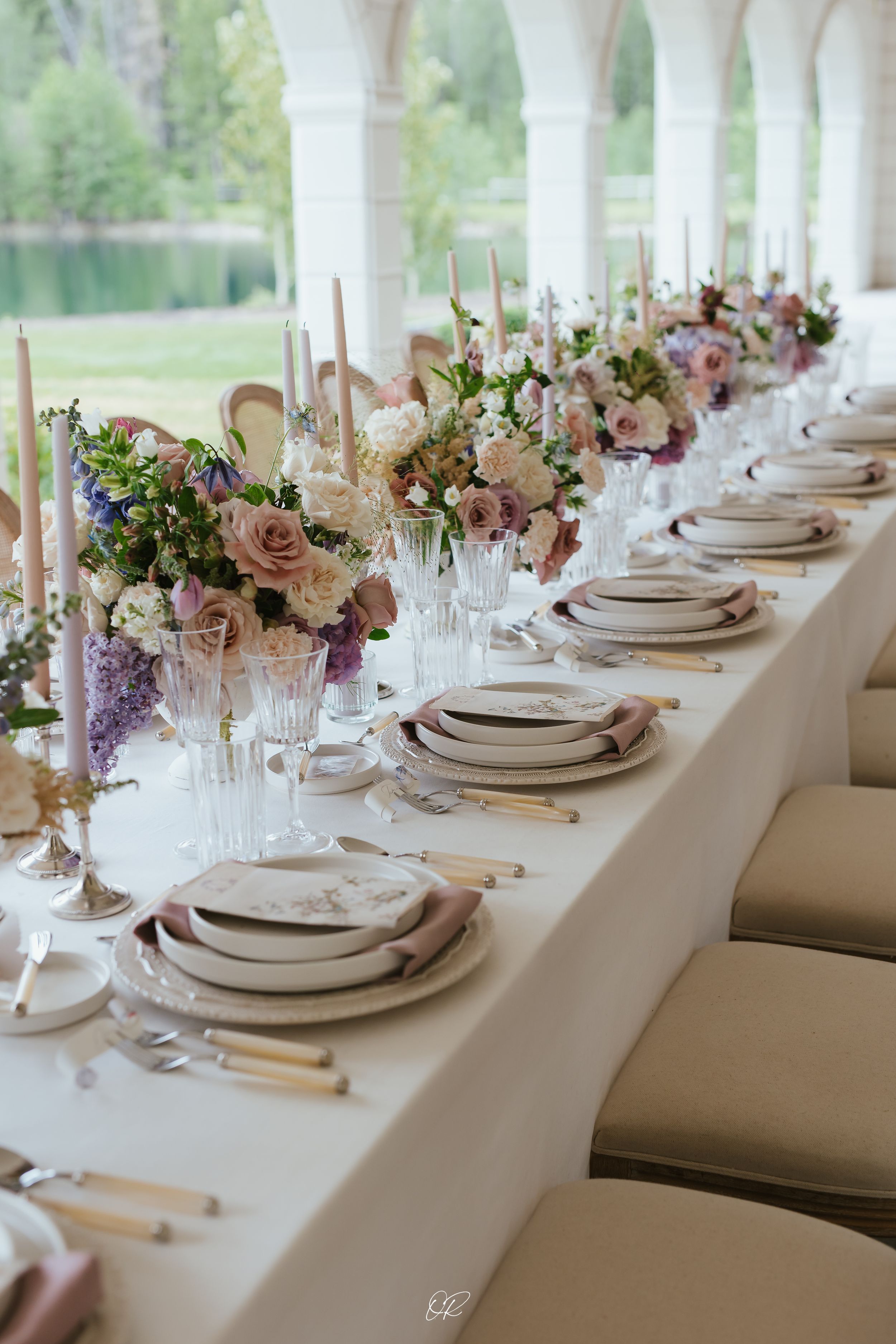 Elegant wedding table setting with pink and purple floral centerpieces, white linens, and formal place settings at an outdoor venue.