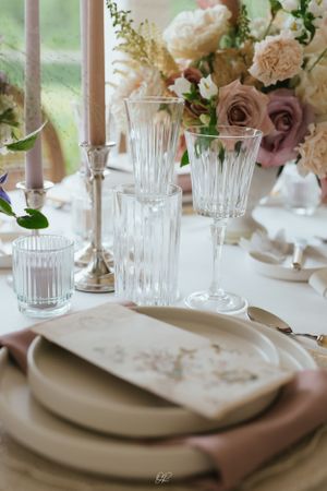 Elegant table setting with crystal glasses, taupe linens, and romantic floral centerpiece by a sunlit window.