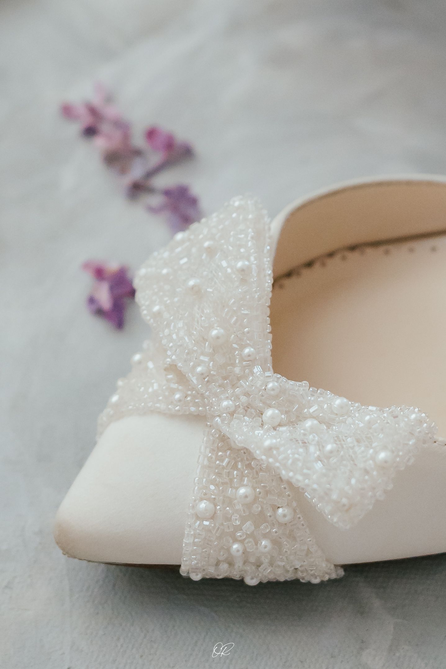 Elegant white bridal flat shoe decorated with a sparkling sequined bow and purple flowers in background.
