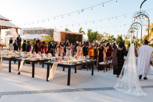 Outdoor wedding reception setup with string lights and long tables at a tropical venue during sunset.