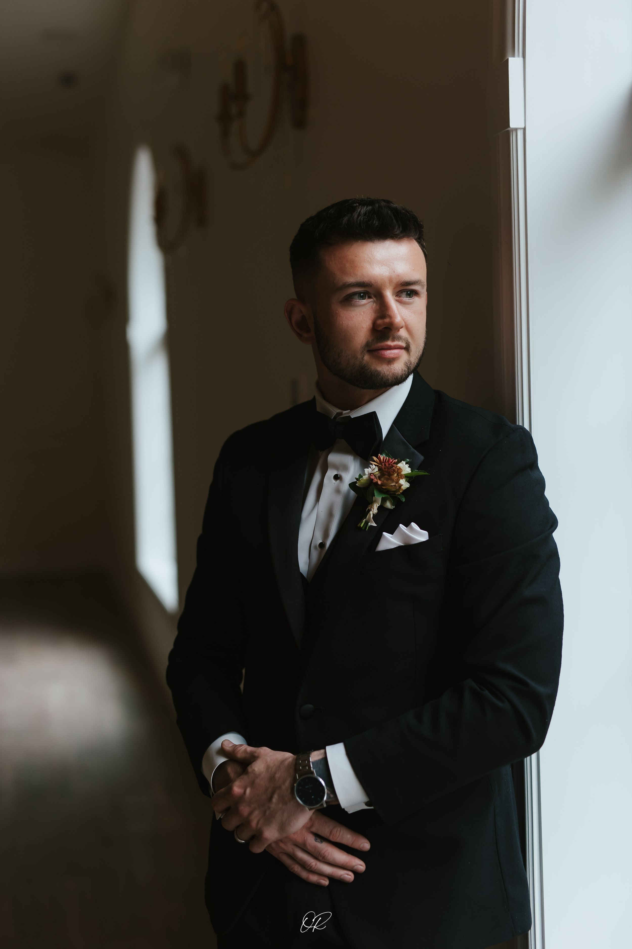 A moody portrait in black and white tones showing a groom in classic tuxedo standing by window light.