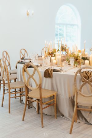 Elegant wedding reception table with wooden cross-back chairs, white linens, and glowing candlelight.