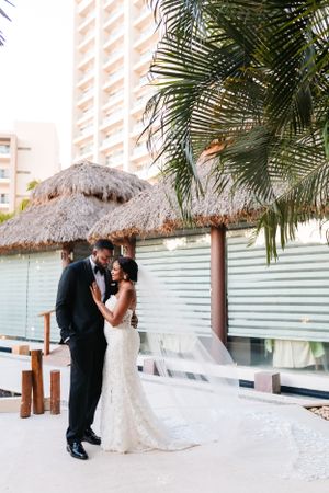 A couple shares a romantic moment under a thatched-roof beach cabana with palm trees and high-rise buildings in the background.