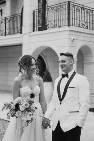 Black and white wedding photo of a couple in formal attire with the bride holding a floral bouquet outside a grand building.