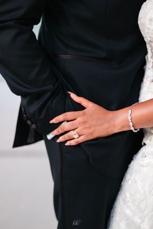A close-up of a bride's hand with a diamond ring and bracelet resting against a groom's black suit.