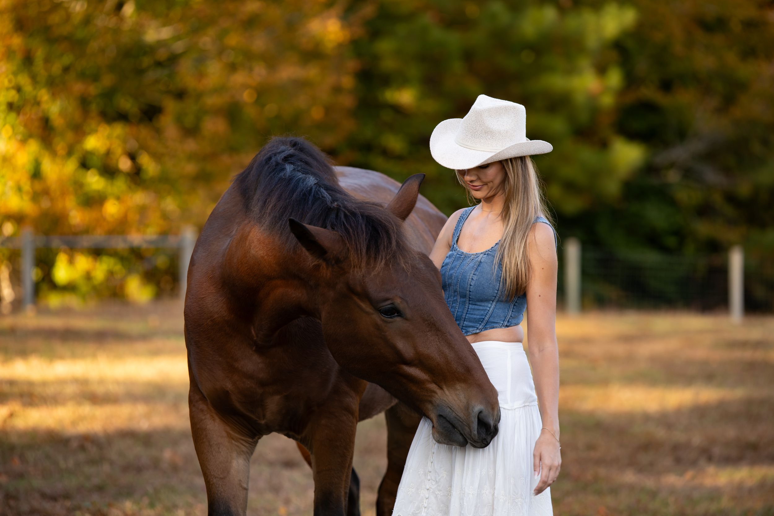A beautiful bay horse stands next to someone wearing a white cowboy hat and long skirt in golden sunset lighting.