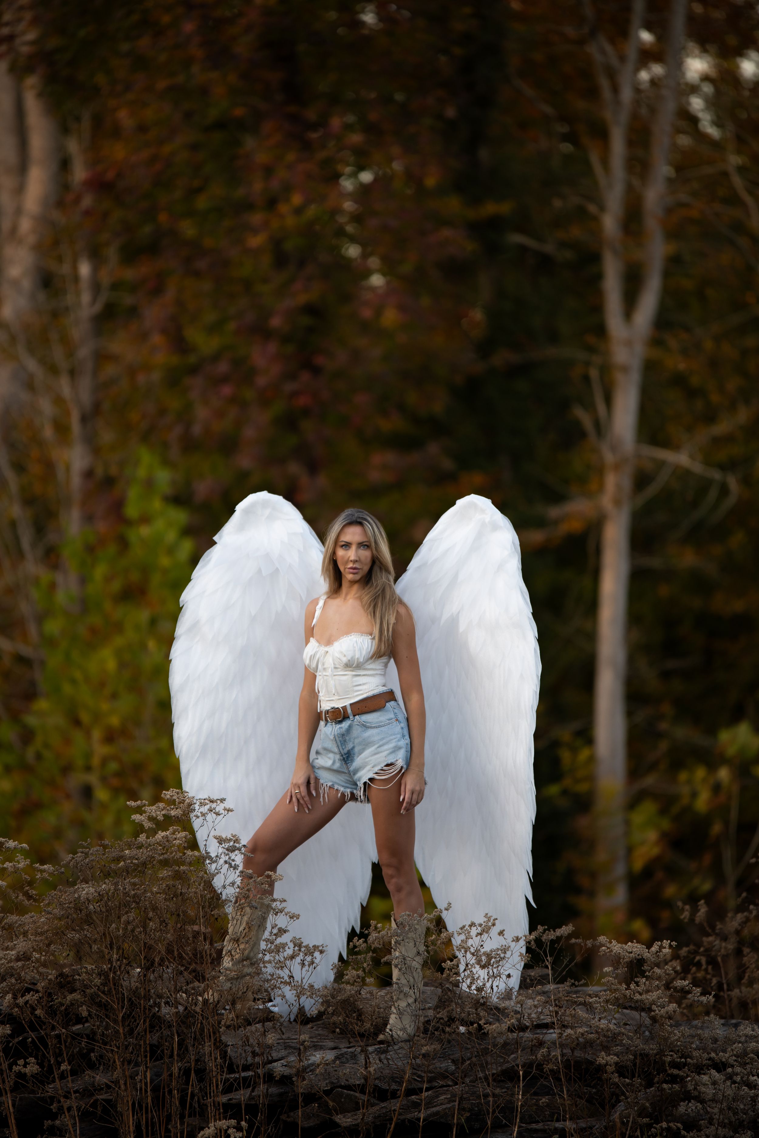 Figure wearing large white feathered wings and denim shorts poses outdoors against autumn forest background.