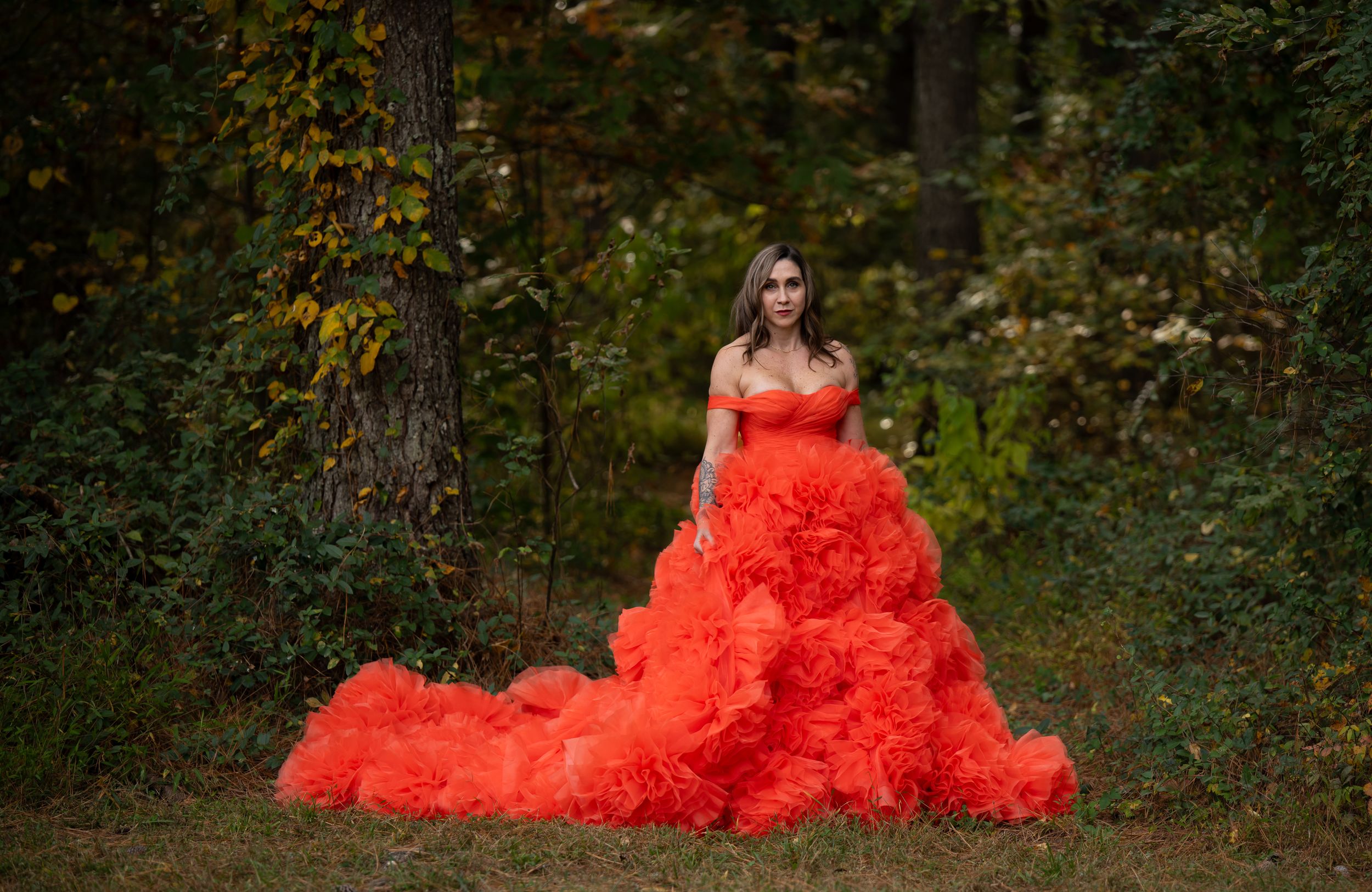 A flowing orange gown with ruffled tulle skirt stands out dramatically against a wooded autumn backdrop.