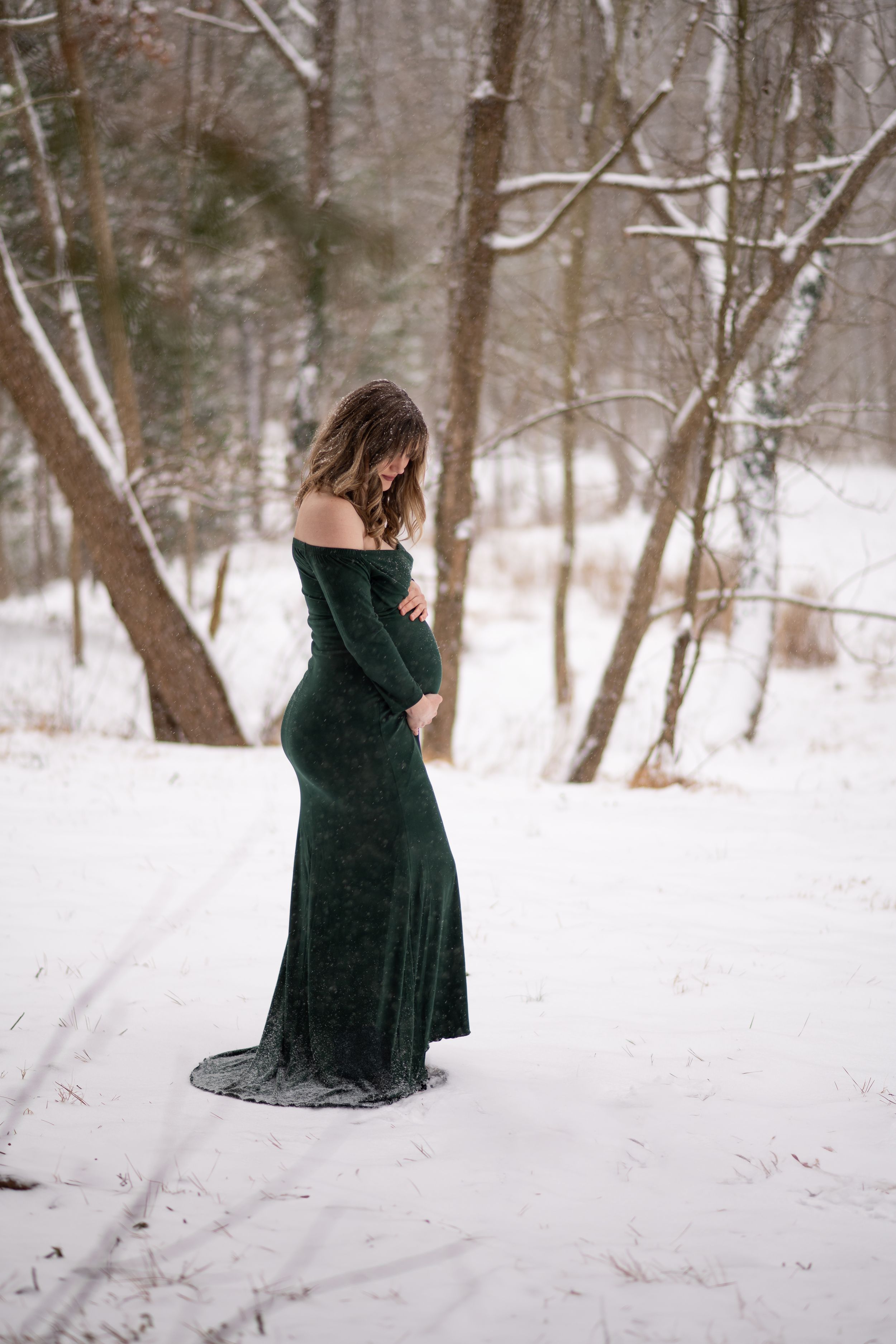 A woman in a long dark velvet dress stands in a snowy forest setting during winter.