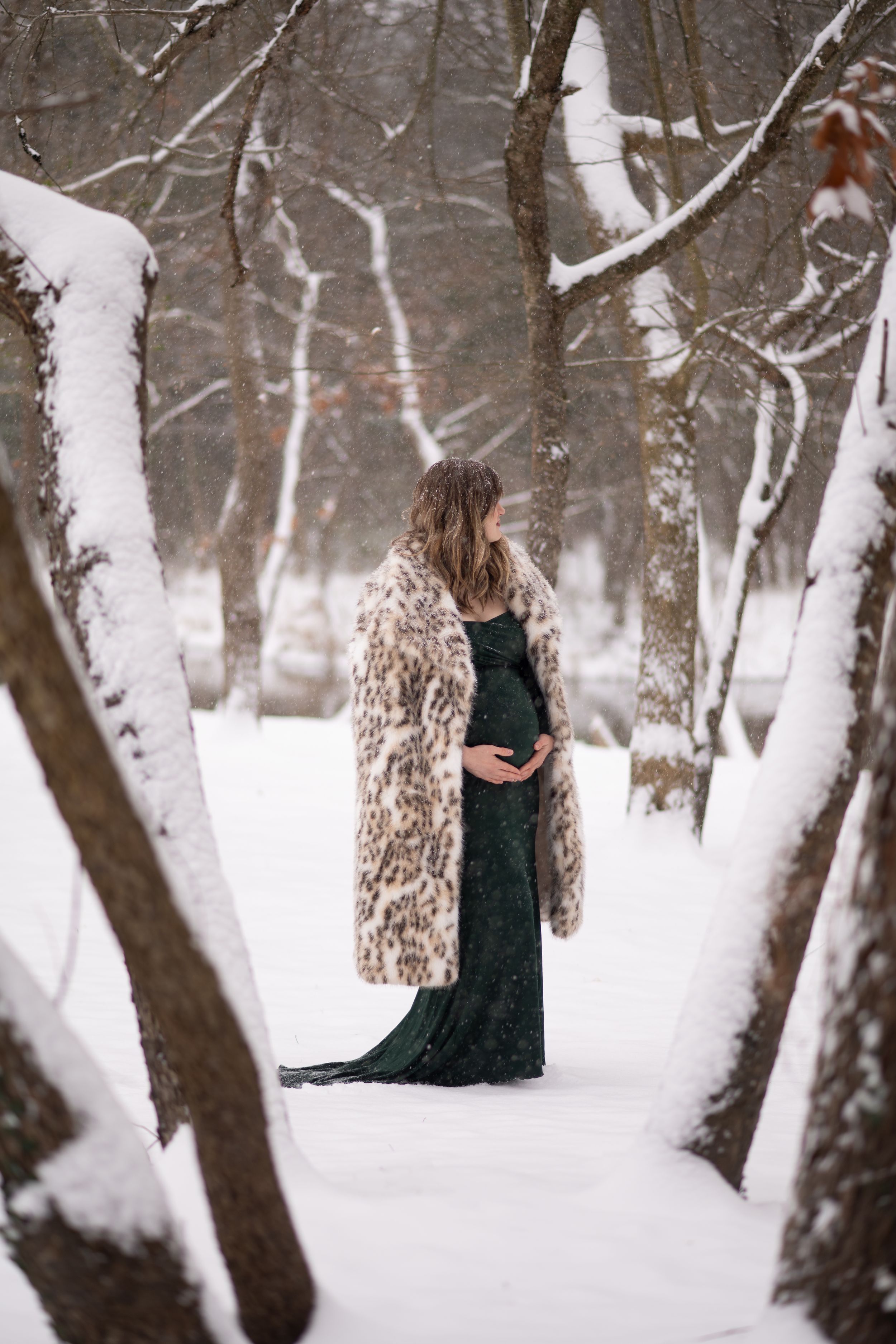 Person in emerald green dress and fur coat stands thoughtfully in snow-covered winter forest path.