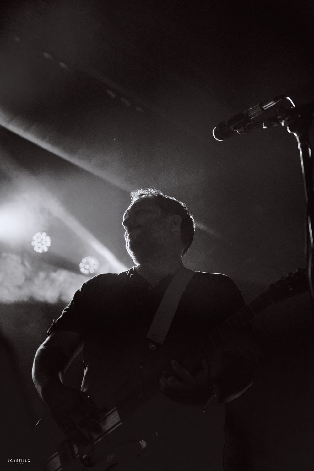 Silhouetted musician playing guitar against bright stage lights in moody black and white composition.