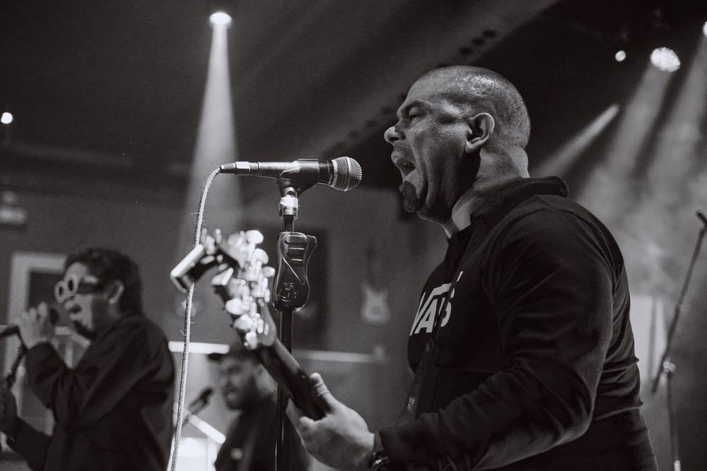 Intense monochrome performance photo of musicians singing into microphones under spotlight.