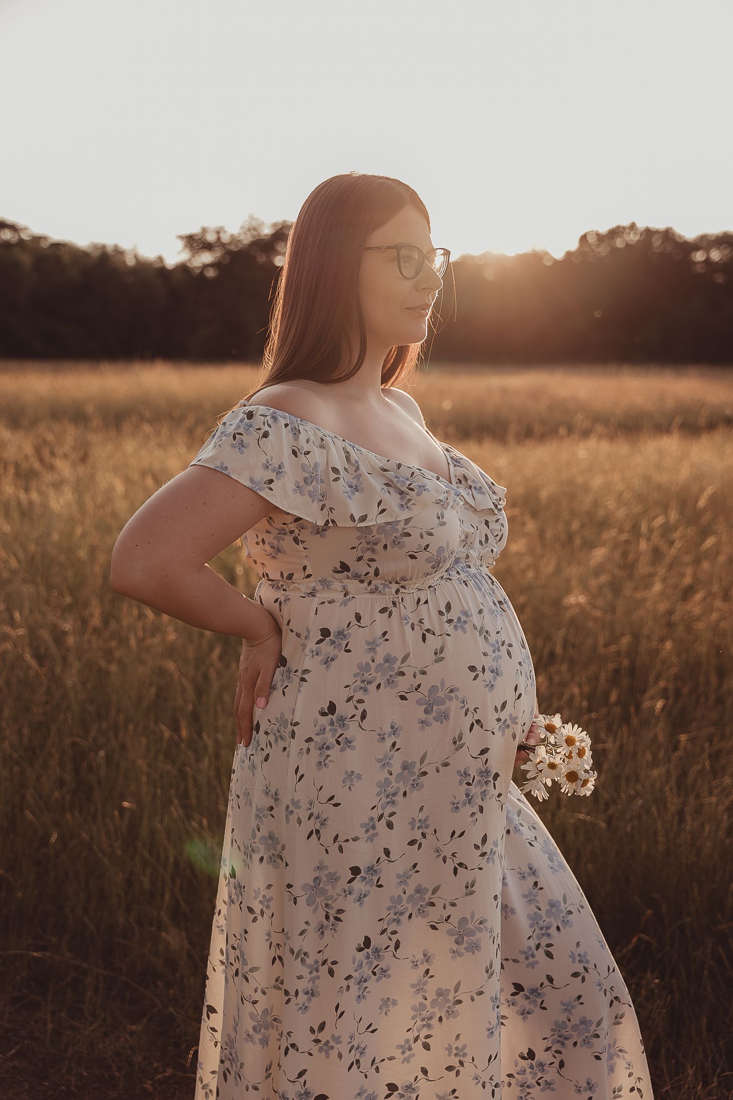 Person wearing a floral dress stands among tall grass in a sun-drenched meadow during golden hour.