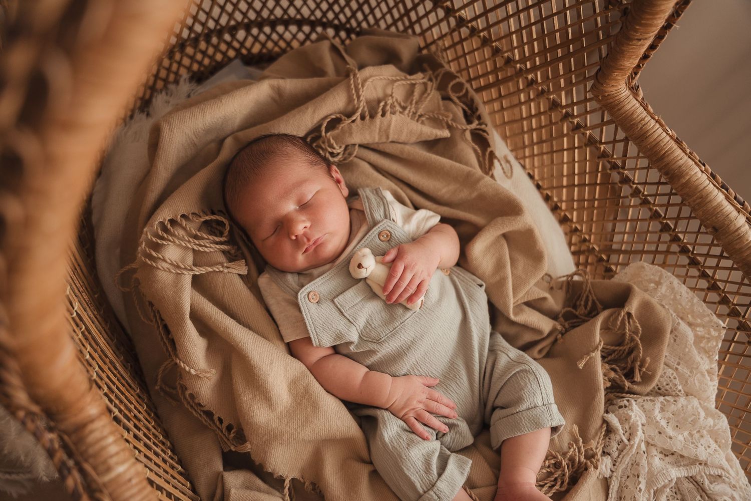 A sleeping newborn baby is peacefully nestled in a cozy brown wicker bassinet with soft beige bedding and lace details.