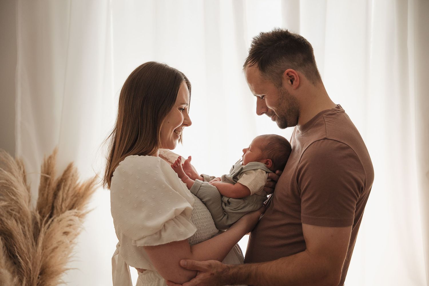 Two people in casual attire lovingly hold their newborn baby near a window with pampas grass decor.
