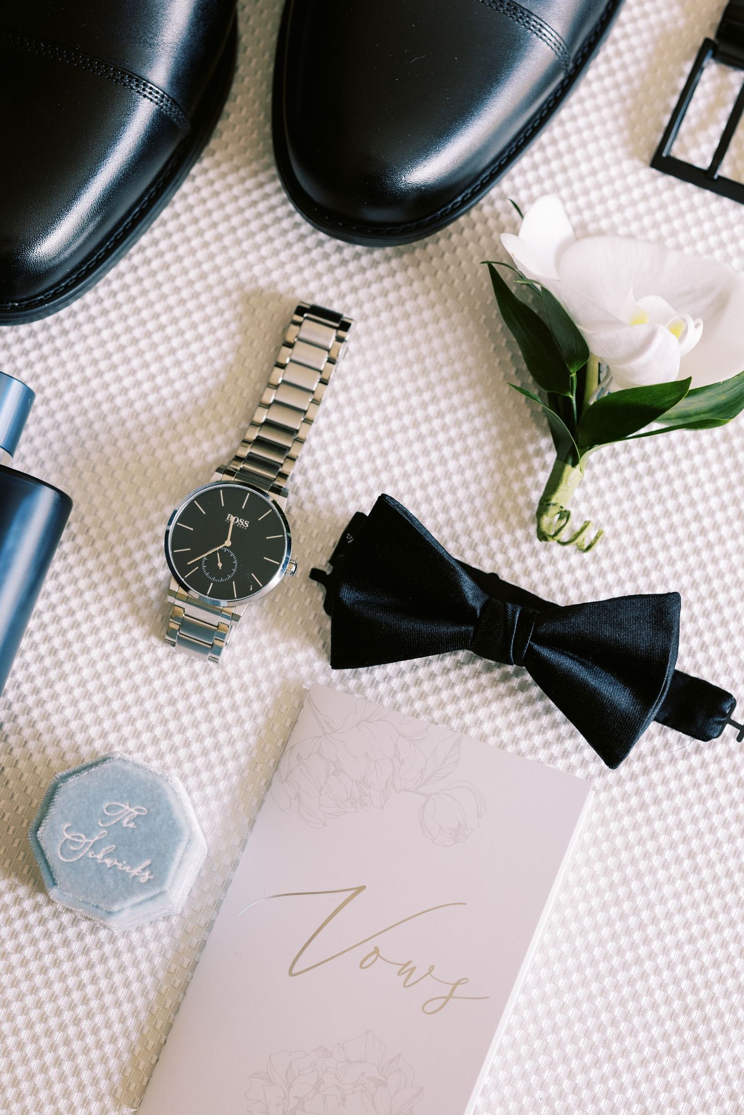 Flat lay of groom wedding accessories including black shoes, bow tie, watch, vows book and boutonniere.
