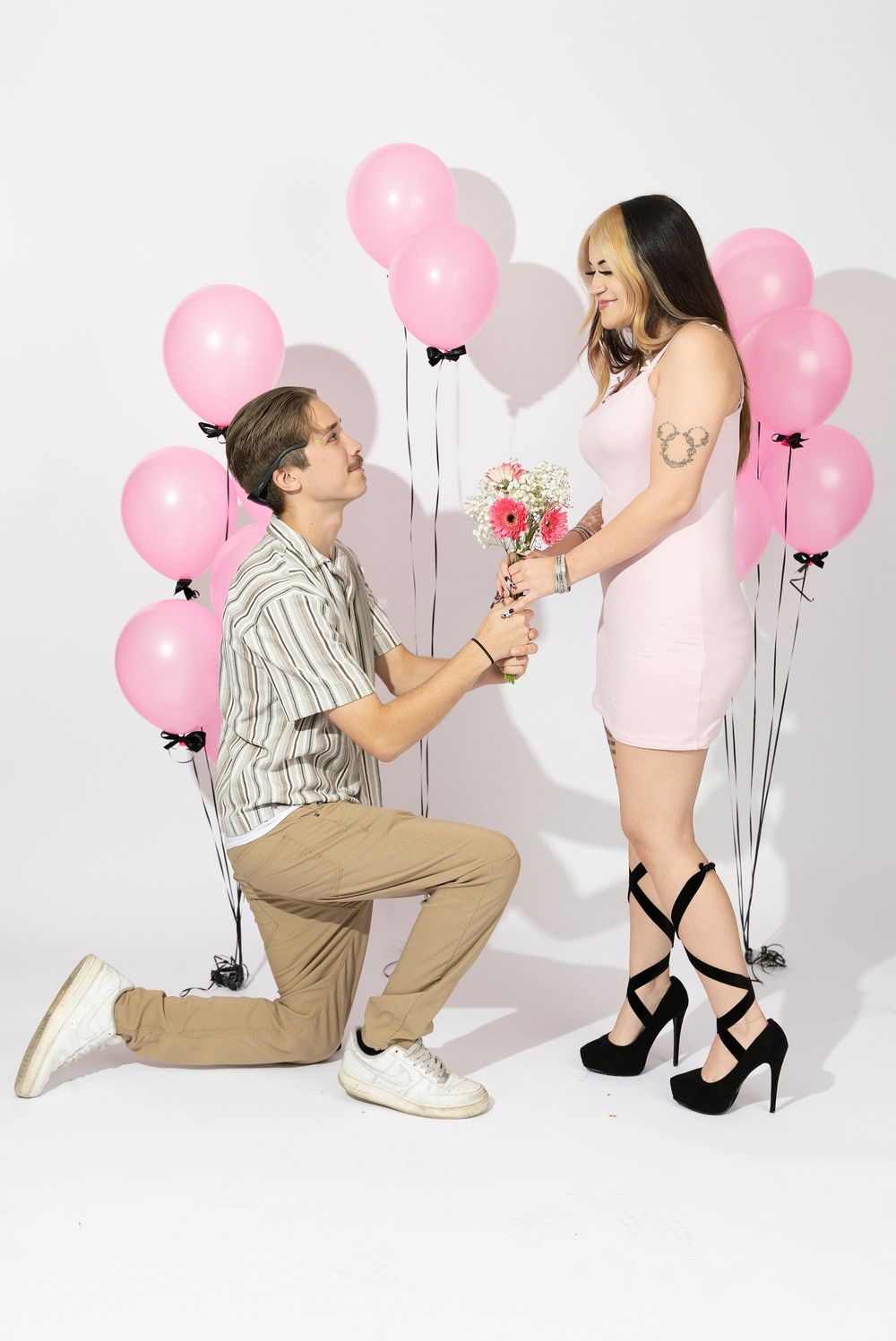 Romantic proposal scene with pink balloons against white backdrop featuring couple in pink dress and khaki pants.