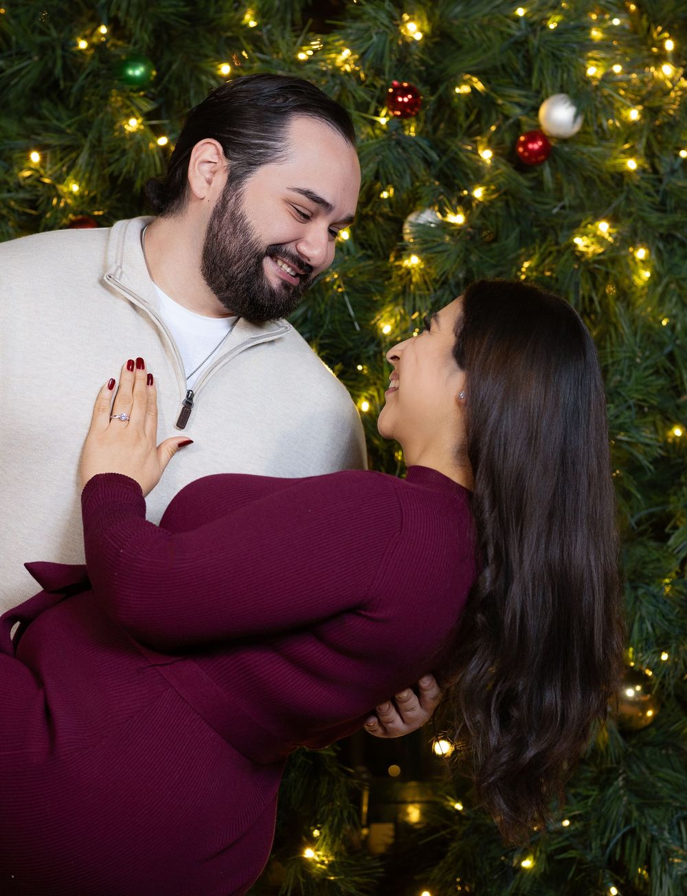 A couple shares an intimate moment in front of a Christmas tree decorated with twinkling lights and ornaments.