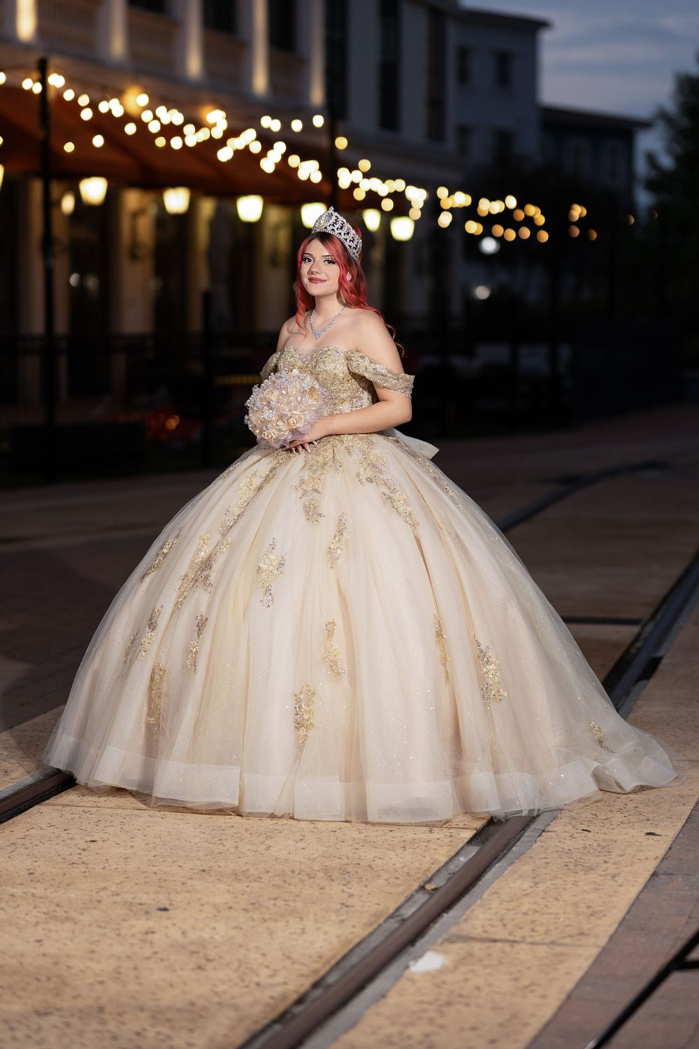 A stunning ball gown in cream with gold embellishments sparkles under evening lights near train tracks.