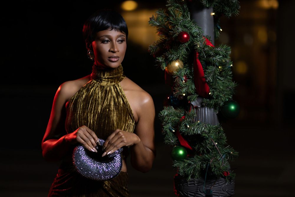 A person in festive holiday attire poses with a decorated Christmas tree and sparkly ornaments.