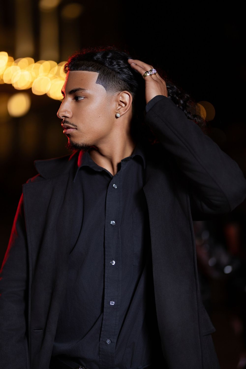 A person in a black button-up shirt adjusts their collar in a dimly lit setting with bokeh lights in the background.