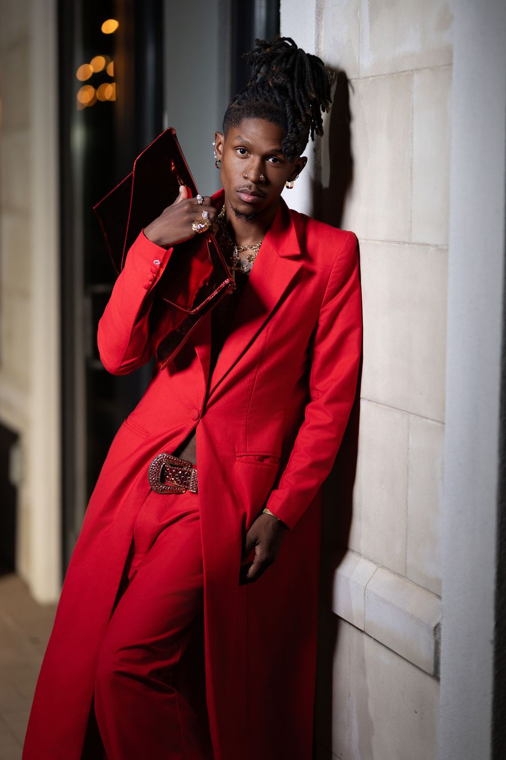 Fashion model in bright red suit jacket poses dramatically against white wall in moody lighting.