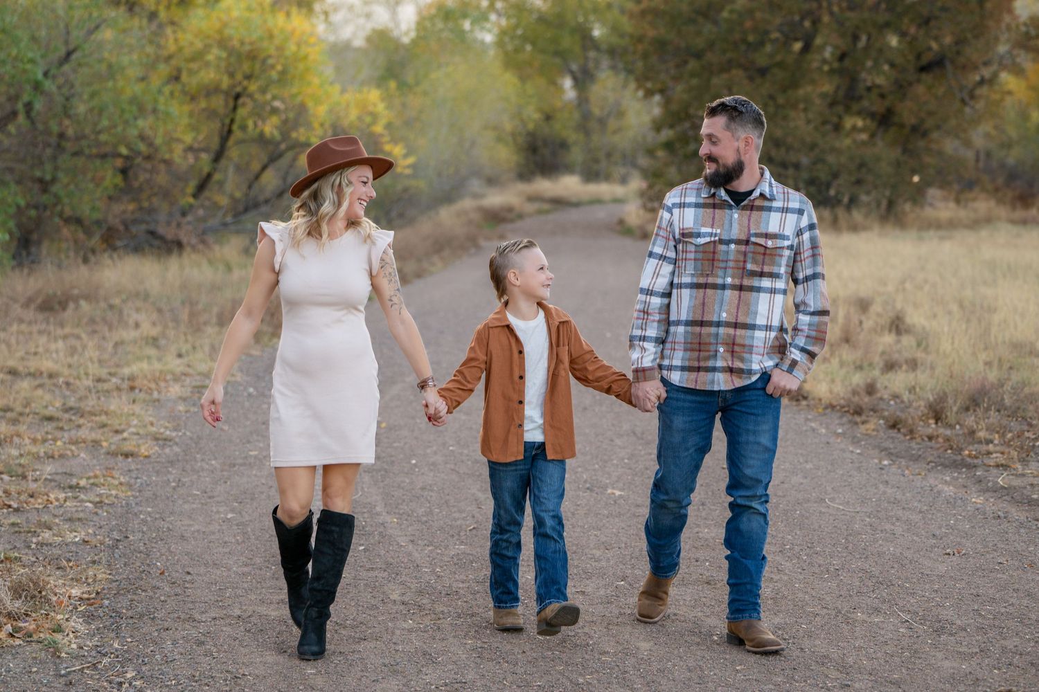 Family of three walking hand in hand on dirt path in autumn setting.