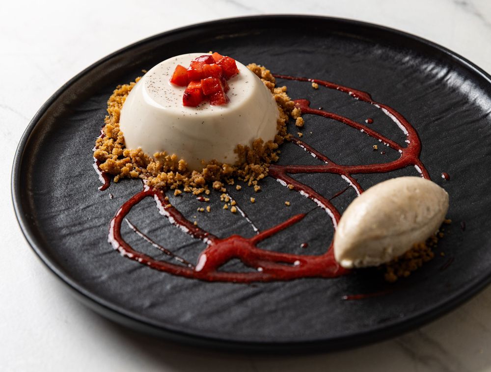 Food photograph of vanilla panna cotta served with a crumble and fresh strawberries served on a black plate with ice cream quenelle.