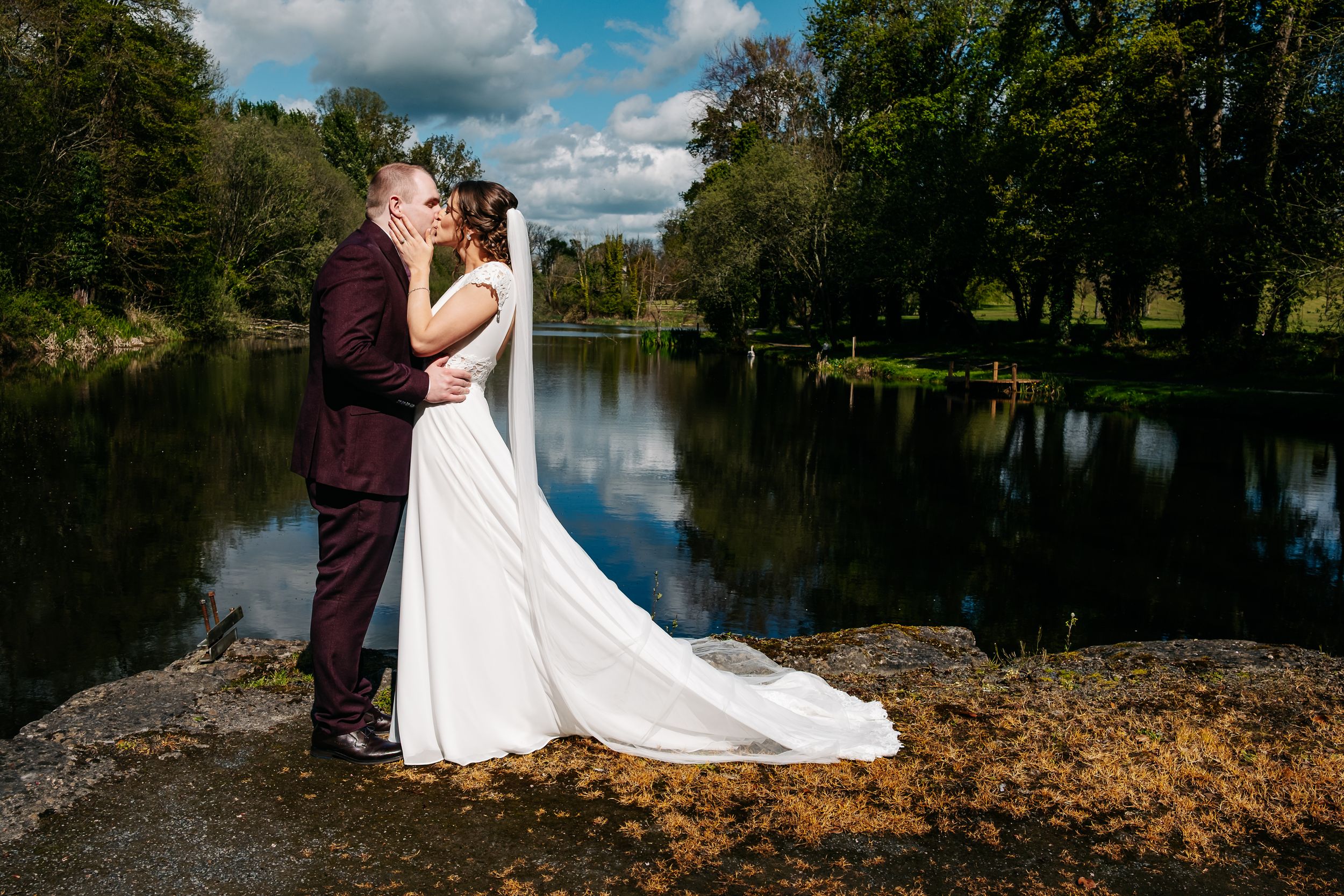 A couple embraces on a riverbank surrounded by autumn trees with a serene lake reflecting the cloudy sky.