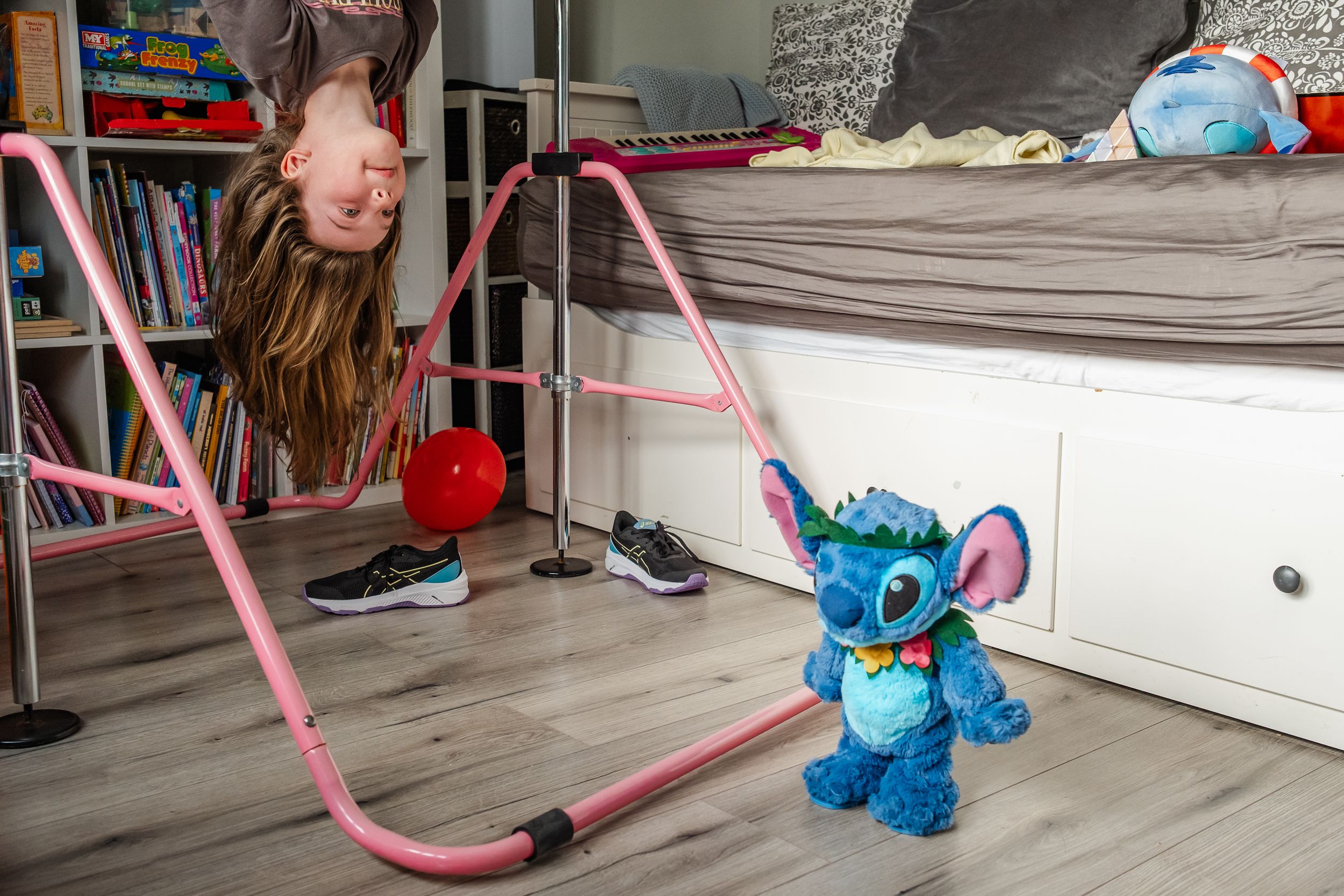 A child hangs upside down from a bed near a blue Stitch plush toy and pink jump rope on the floor.