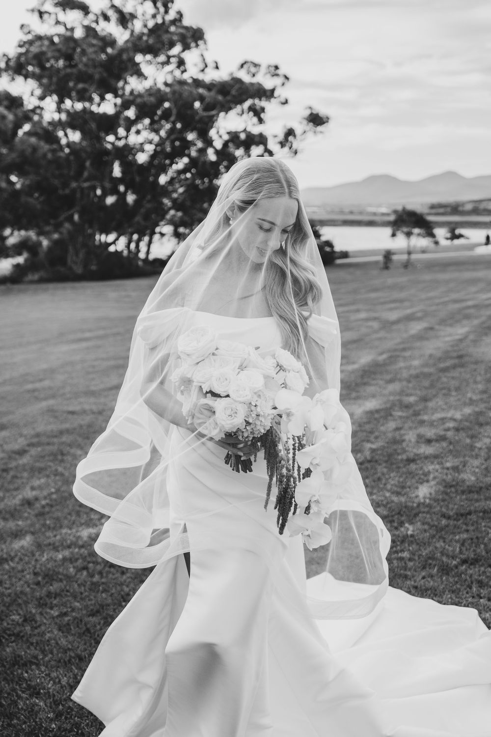 Black and white photo of a bride in a flowing gown and veil, holding a bouquet, standing on a lawn with mountains behind.