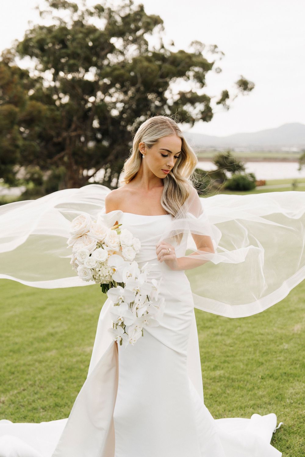 Blonde bride in white off-shoulder gown holds floral bouquet as veil flows in breeze on lush green lawn.