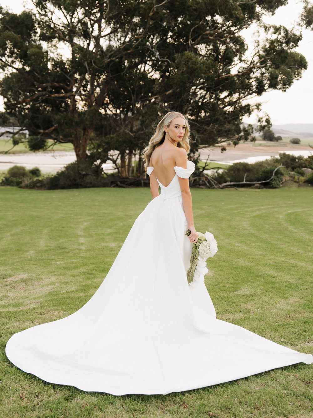 Bride in off-shoulder white ball gown with long train poses on green lawn with trees in background.