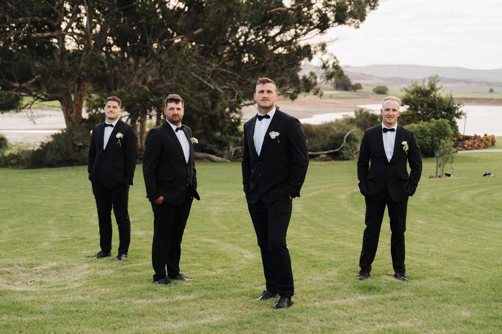 Four groomsmen in black tuxedos with bow ties stand on green grass with scenic coastal landscape behind them.