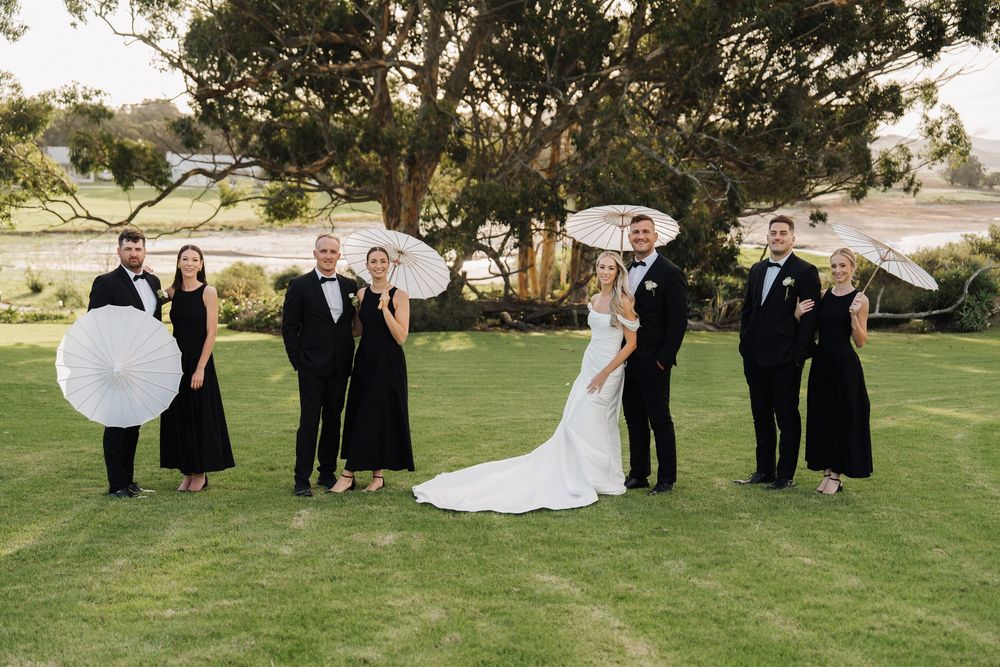 Wedding party in black attire holding white parasols on green lawn with trees.