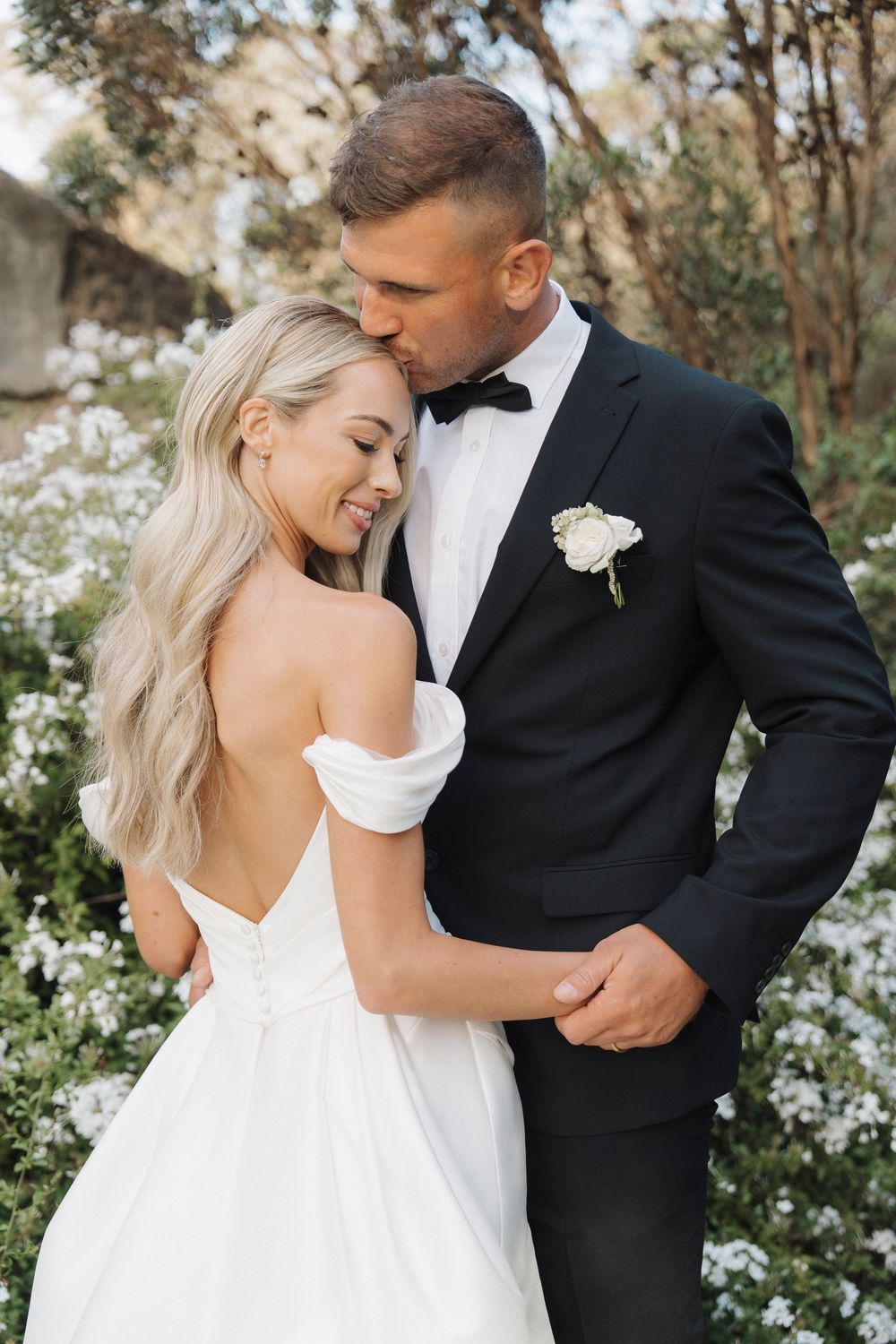 Groom in black tuxedo kissing blonde bride in white off-shoulder gown outdoors.