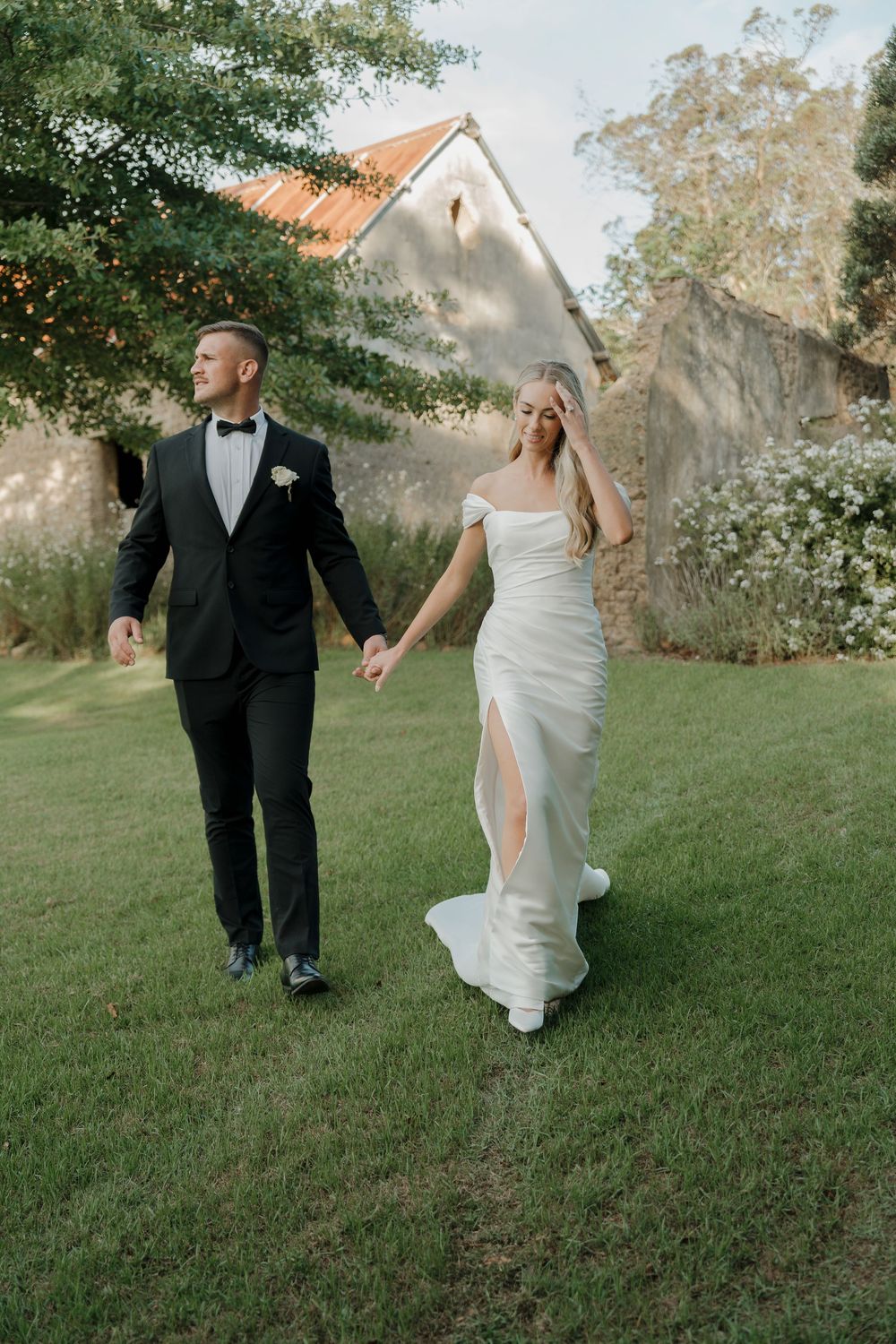 Bride and groom walk hand in hand across green lawn near rustic stone building, bride in white off-shoulder gown.