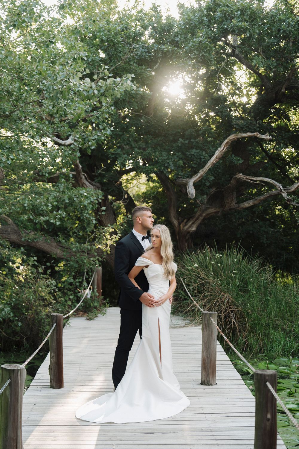 Bride and groom embracing on wooden bridge surrounded by lush green trees in golden sunlight.