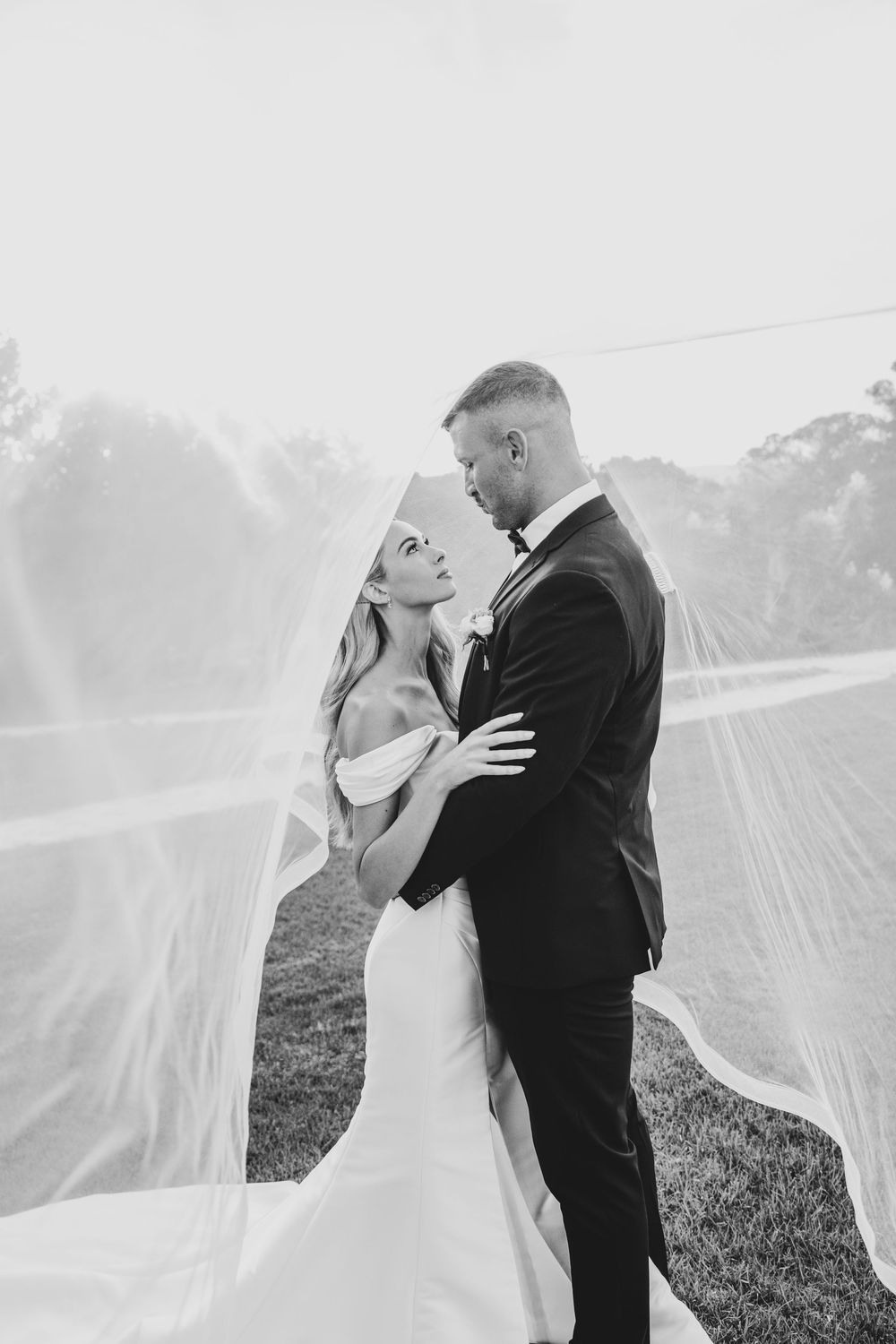 Black and white photo of bride and groom embracing under flowing veil outdoors on their wedding day.
