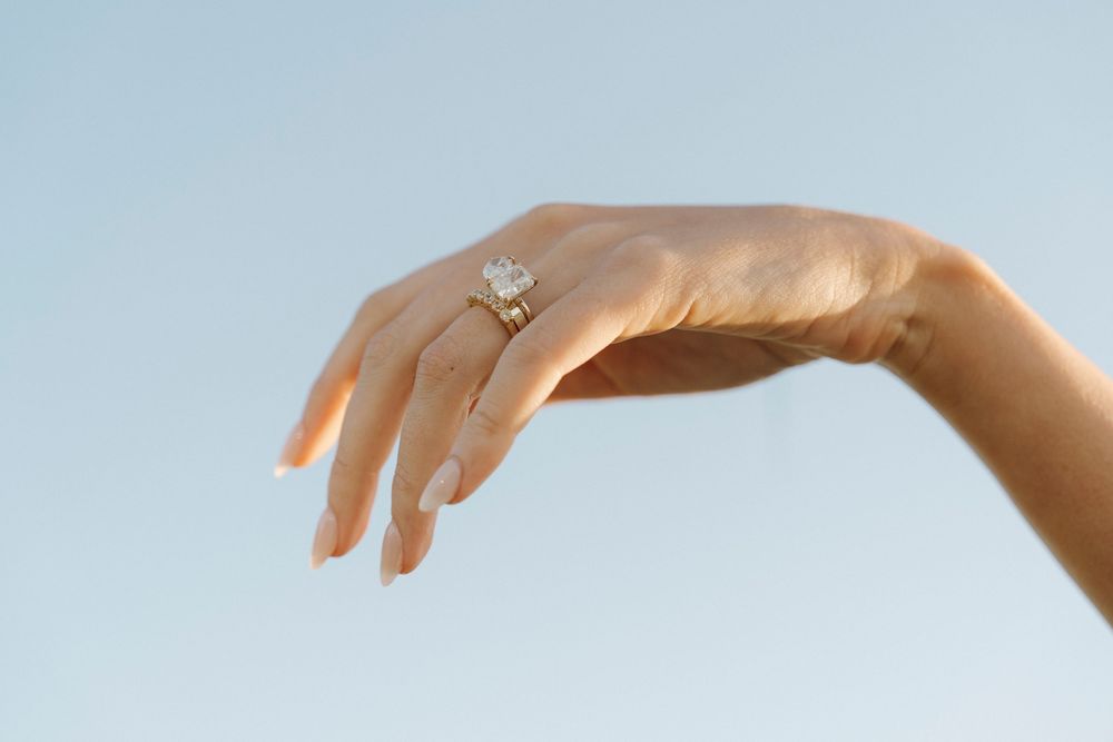 A woman's hand elegantly raised against a blue sky, wearing a stunning gold diamond engagement ring.