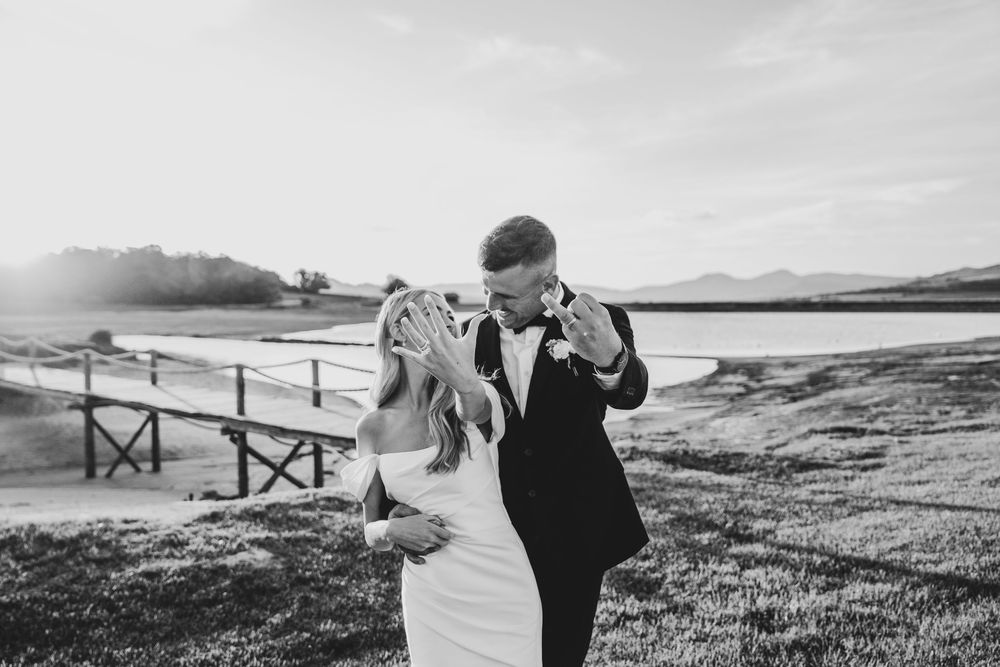 Black and white photo of a bride and groom sharing a tender moment outdoors near a lake on their wedding day.