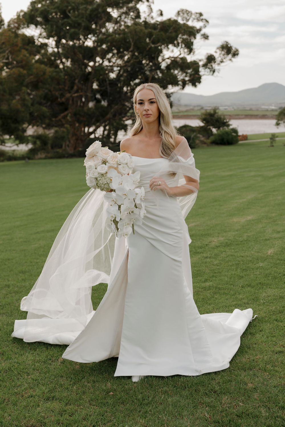 Bride in off-shoulder white gown holding cascading white floral bouquet on green lawn with mountain backdrop.