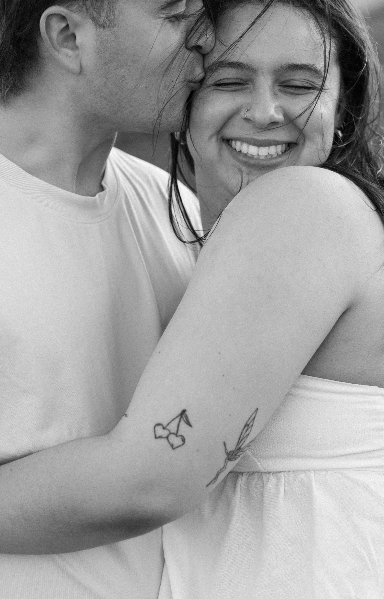 A smiling woman with tattoos on her arm is embraced by a man in a black and white couples photo.