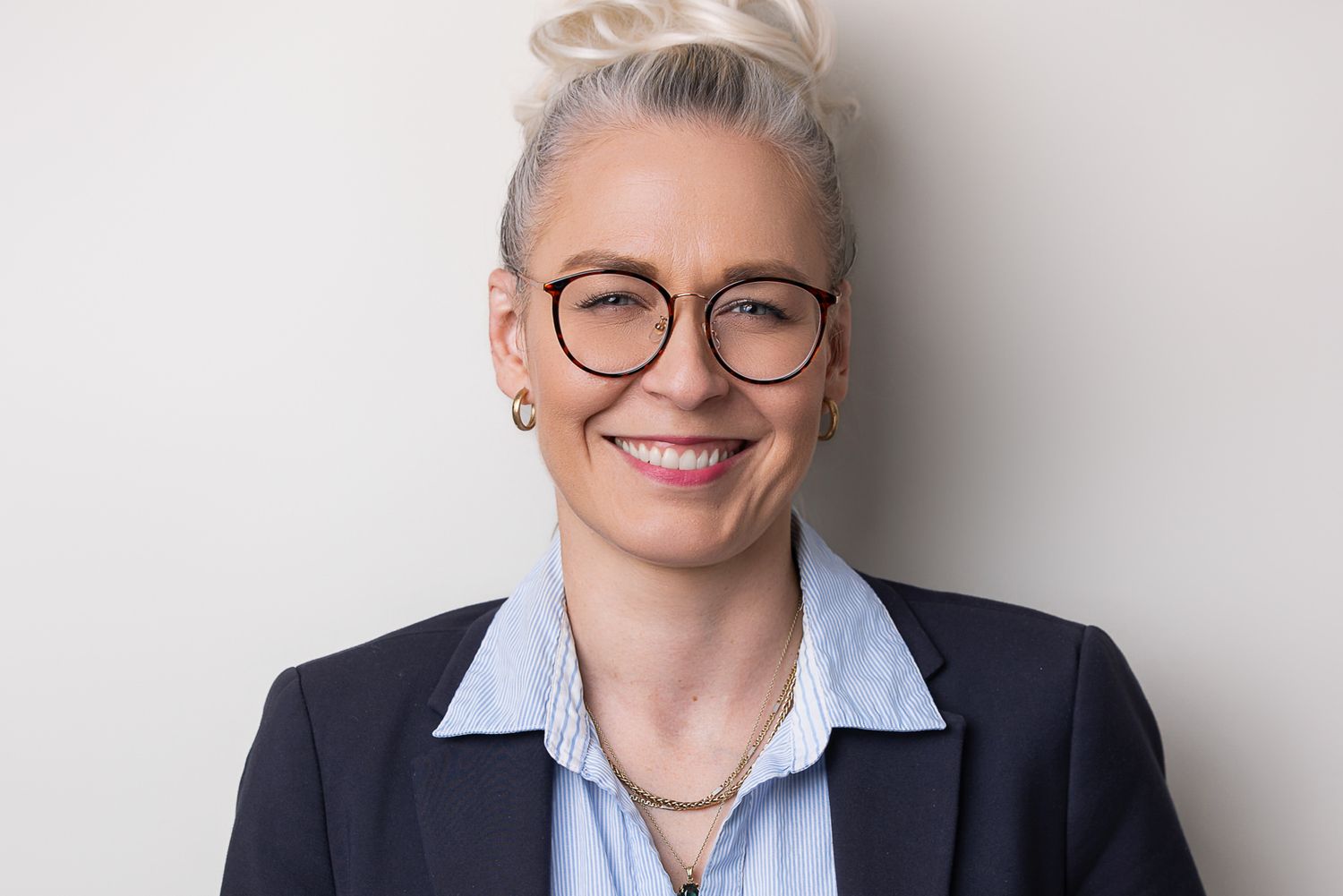 Professionelles Studioporträt einer Person mit braunem Blazer und schwarzem Rollkragenpullover vor dunklem Hintergrund.