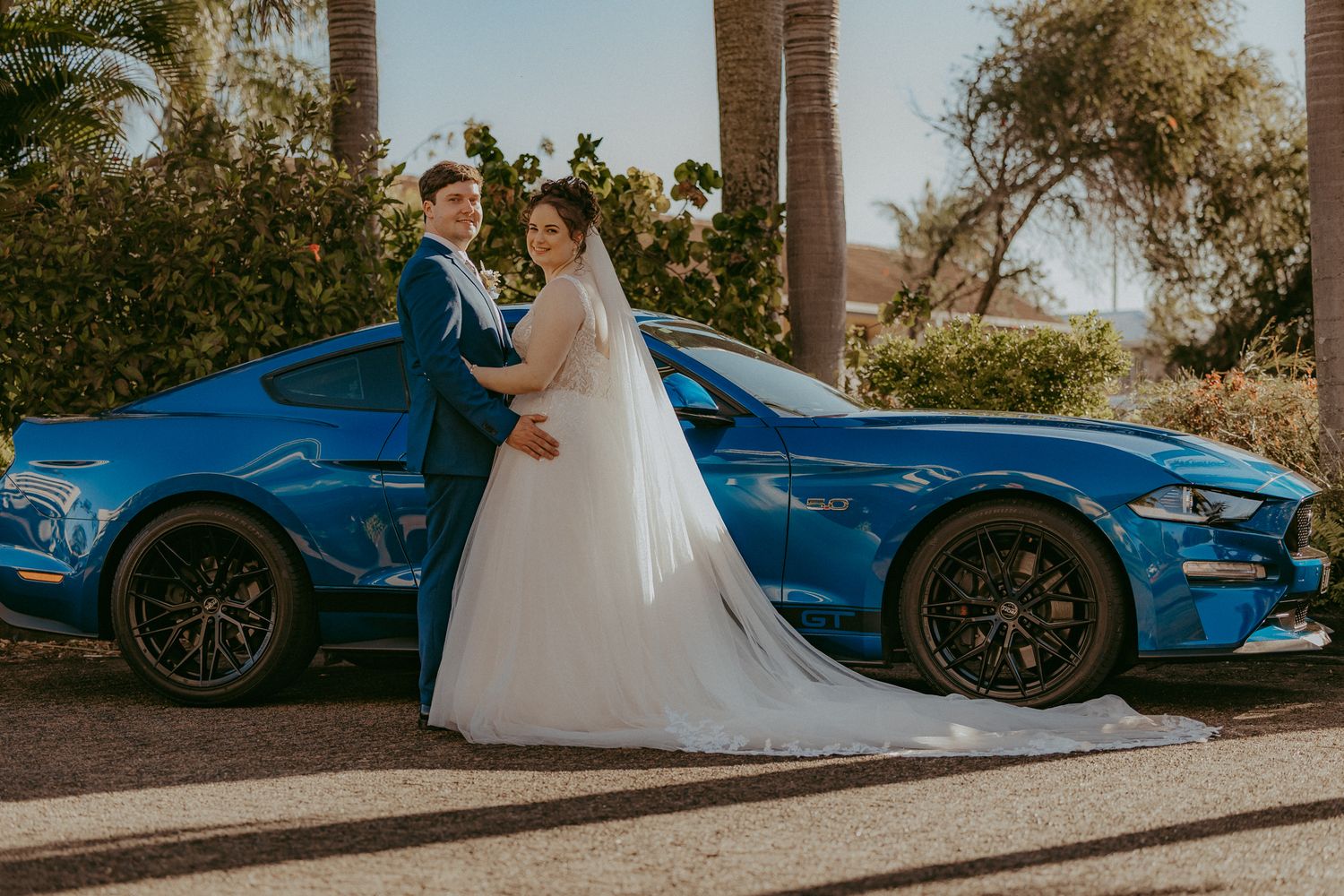 Bride and groom pose together in front of a blue Ford Mustang at their wedding venue.